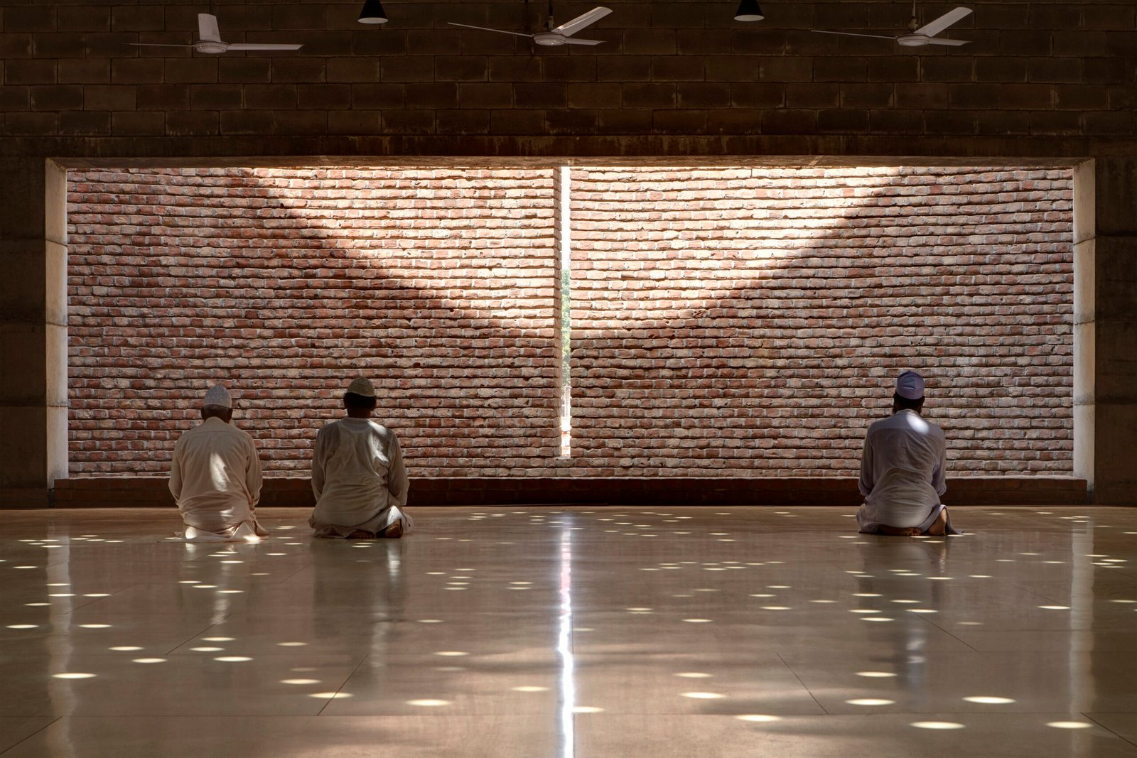 Three worshippers kneel facing the qibla wall, where a vertical slit of light marks the direction of Mecca, circular light patterns scattered across the polished floor