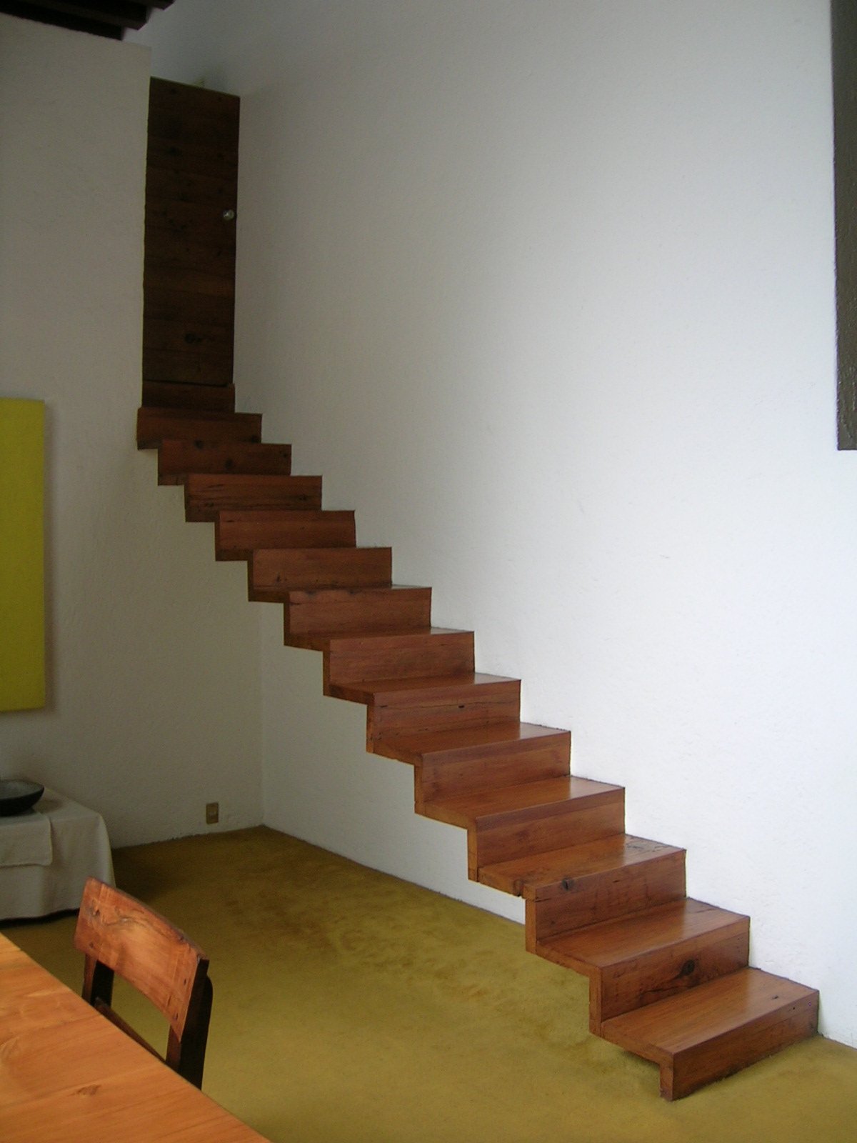 The cantilevered wooden staircase in the library of Casa Luis Barragán, solid treads rising without a railing against white plaster, a yellow painting visible at left