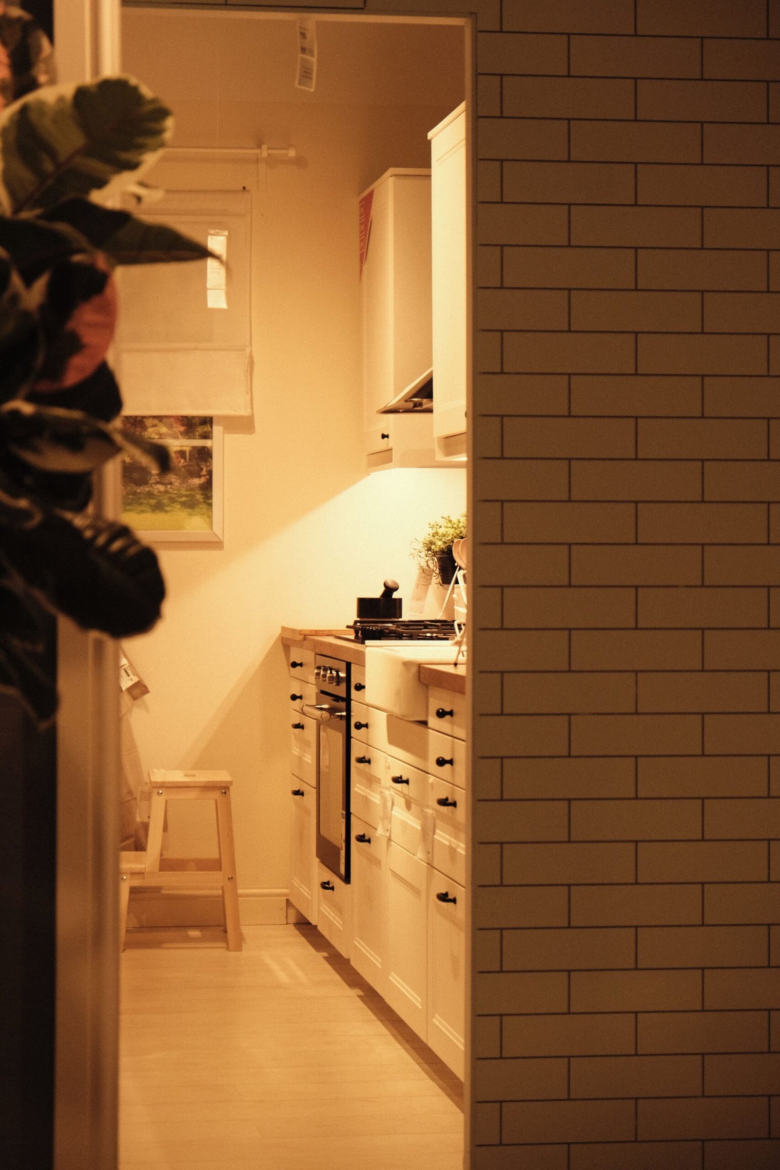 A narrow galley kitchen seen through a doorway, white cabinets and a wooden step stool under warm light.