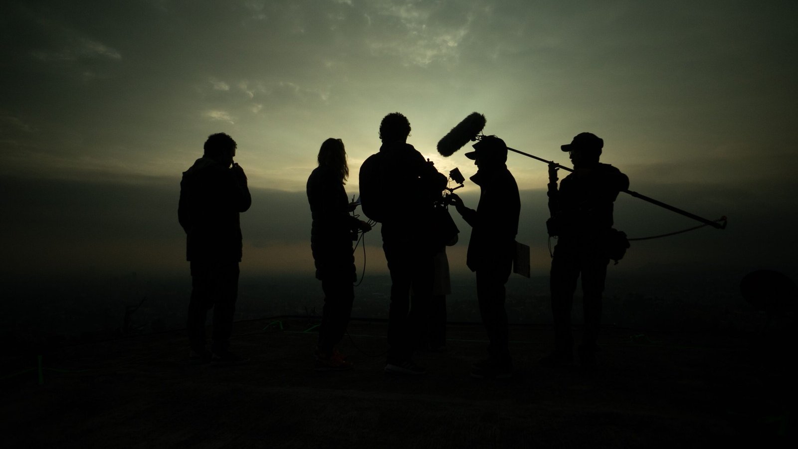 A documentary film crew in silhouette against a low evening sky, a boom microphone raised over the group.